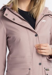Veste imperméable rose clair avec capuche, ferrures argentées et fermeture à boutons sur le devant. Présente un logo minimaliste de parapluie sur la poitrine.