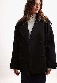 Cappotto doppiopetto nero con ampi rever, tessuto testurizzato e dettagli con bottoni. Indossato sopra un maglione a collo alto grigio chiaro.