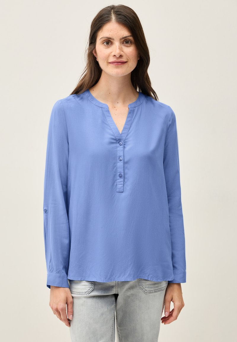 Camicia a maniche lunghe azzurro chiaro con scollo a V e tre bottoni, con una vestibilità rilassata e una texture morbida. Indossata sopra dei jeans grigi chiari.