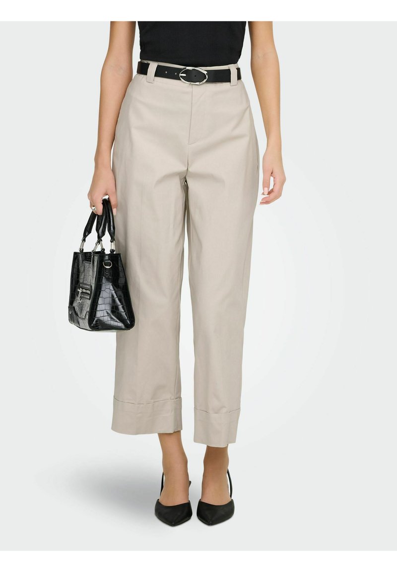 Femme portant un pantalon beige taille haute et court, une ceinture noire, un haut noir, des talons noirs pointus, et tenant un sac à main noir texturé.