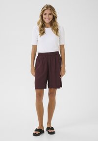 Shorts bordeaux avec une taille élastique, deux poches latérales et une coupe ample, associés à un haut blanc à manches courtes et des sandales noires.