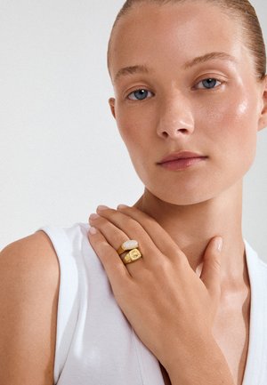 Mujer joven con piel clara con top blanco sin mangas, mano cerca de la clavícula, lleva dos anillos de oro, uno con piedra blanca y otro con grabado de sol.