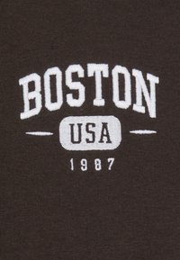 Bruine stof met witte geborduurde tekst: "BOSTON USA" boven "1987." Eenvoudig ontwerp, gladde textuur en geen aanvullende details of patronen.