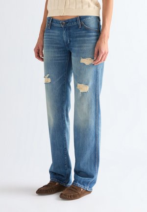 Blaue Jeans mit geradem Bein und abgenutzten Stellen an den Oberschenkeln, kombiniert mit braunen Mokassins und einem beigen, bauchfreien Pullover.