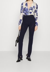 Blusa floreale a maniche lunghe color crema con fiori blu; abbinata a pantaloni attillati blu navy con spacco laterale; stivali neri a punta e borsa.