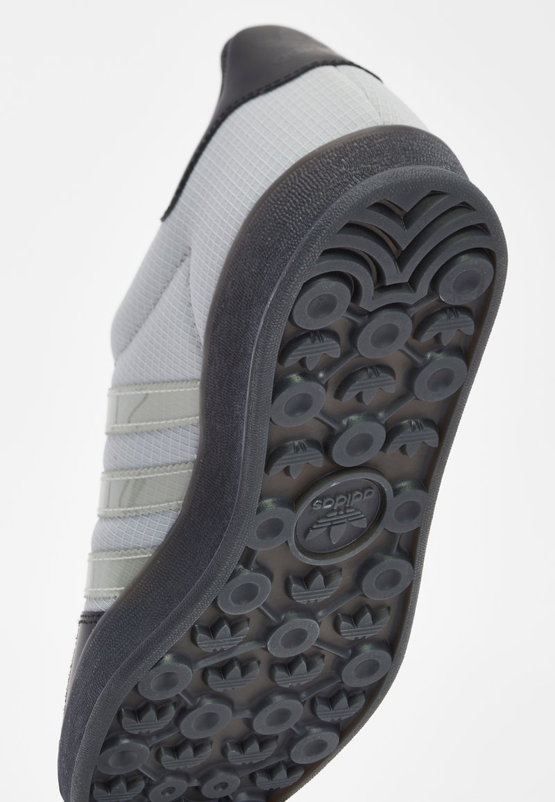Graue Sneaker mit strukturiertem Obermaterial, schwarzen Lederelementen und einer gummierten, gemusterten Außensohle mit runden Griffen und Logos.