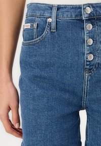 Høyt liv blå denimjeans med fem sølvknapper, synlig søm og en sidelomme med merkeetikett. Glatt tekstur.