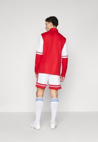 Piros és fehér sportos tréningruha hosszú ujjakkal, csíkokkal az ujjakon és a nadrágon. Fehér zoknik kék csíkokkal és fehér futócipők.
