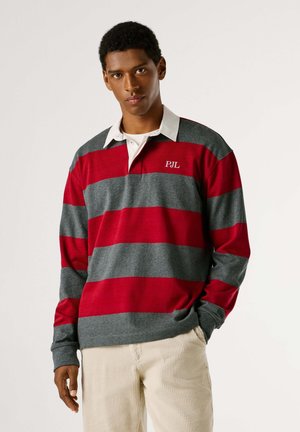 Polo a maniche lunghe a righe rosse e grigie con colletto bianco e logo "PJL" ricamato sul petto, indossato con pantaloni di velluto a coste di colore chiaro.