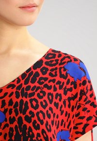 Tejido rojo con estampado de leopardo negro y acentos florales azules; cuenta con un corte holgado y un diseño de escote ancho.
