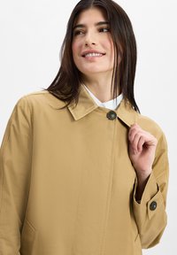 Khaki trench coat with a collar, single button closure, and side pockets. Smooth texture and relaxed fit. Accents include a black button.