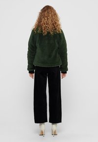 Grön fleecejacka med en texturerad yta, tillsammans med svarta kordbyxor. Outfiten har en avslappnad passform och kontrasterande ljusa skor.