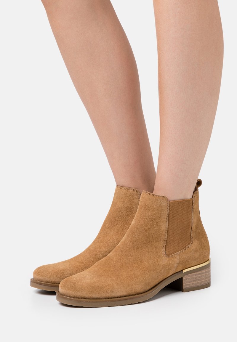 Bottines en daim beige avec talons empilés bas portées sur jambes nues sur un fond clair uni.