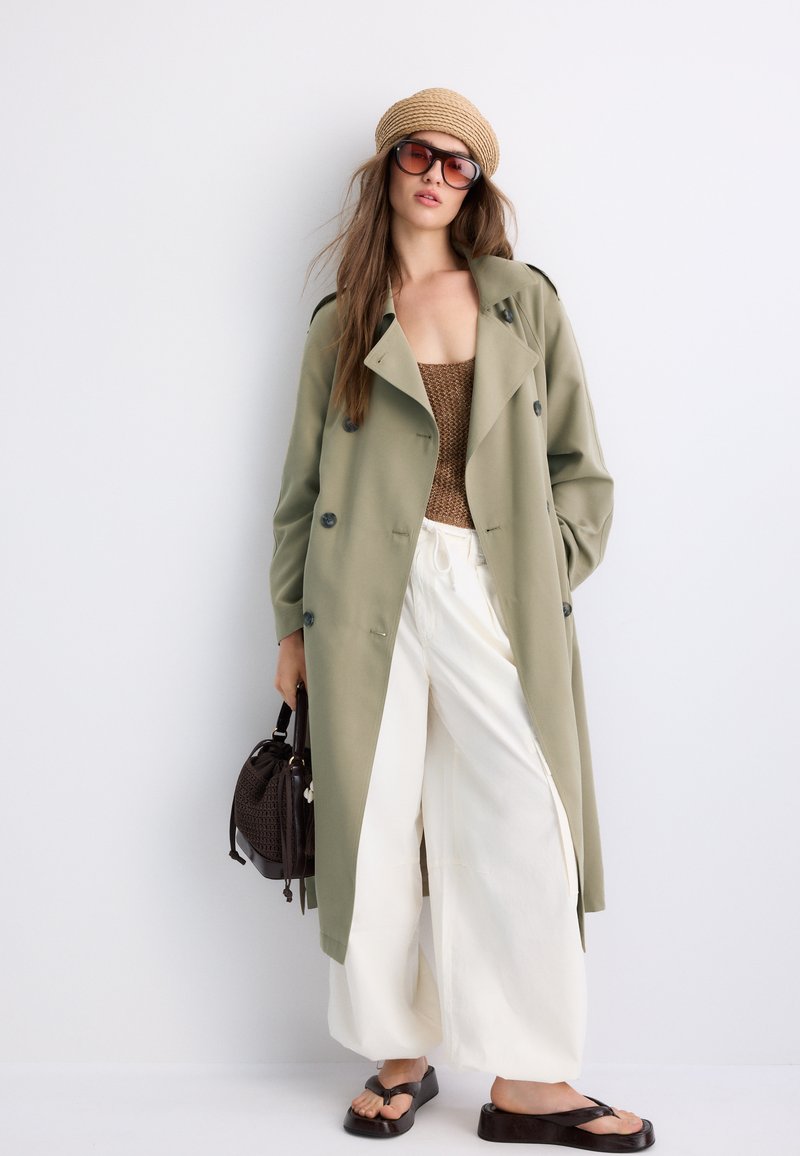 Femme portant un trench-coat olive, un top en maille marron, un pantalon blanc à jambes larges, un chapeau en paille, des lunettes de soleil, tenant un sac à main noir et des sandales à plateforme noires.
