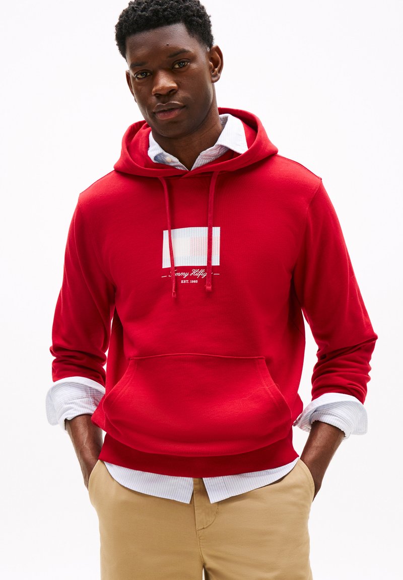 Red hooded sweatshirt with a front pocket and white graphic design. Layered over a white and blue striped shirt. Casual fit, soft texture.