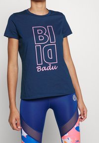 Námořnická modrá bavlněná tričko s odvážným, vertikálním růžovým textem "BID" a "Badu." V kombinaci s vysokým pasem, barevnými legínami s síťovinovými akcenty.