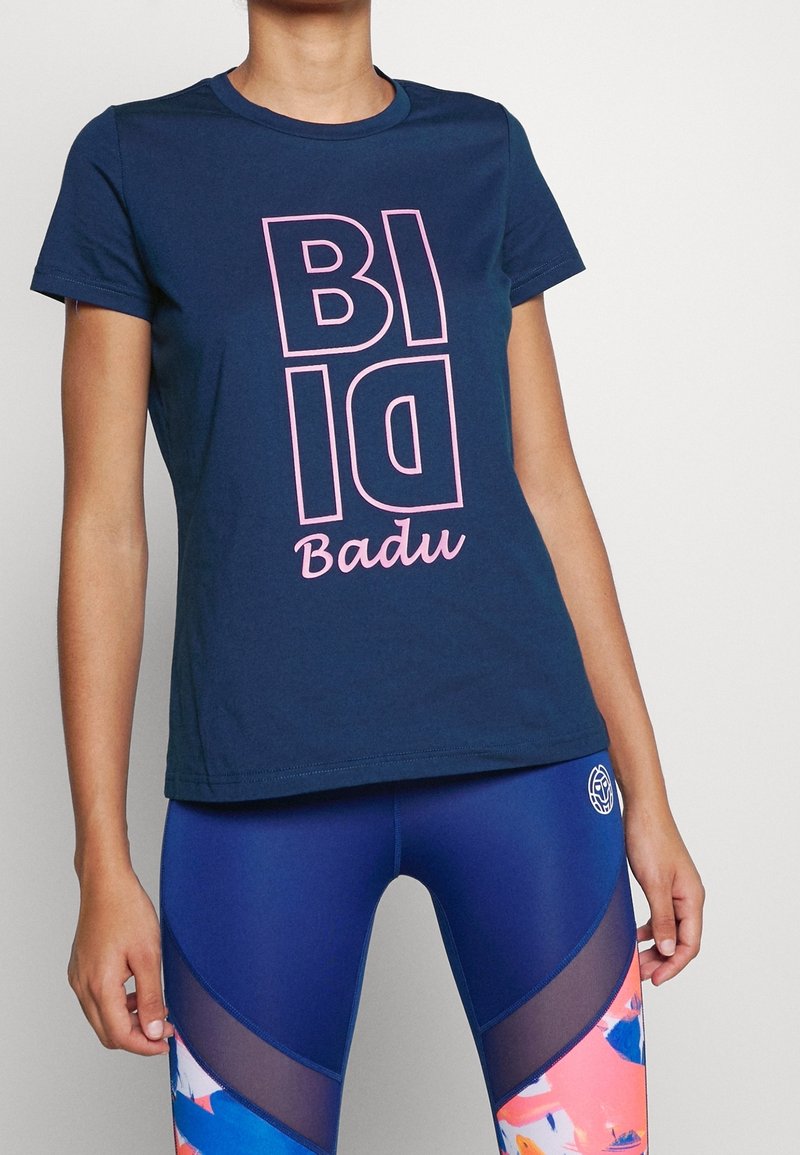 Námořnická modrá bavlněná tričko s odvážným, vertikálním růžovým textem "BID" a "Badu." V kombinaci s vysokým pasem, barevnými legínami s síťovinovými akcenty.