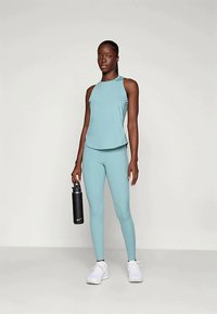 Lyseblå treningssett som inkluderer en ermeløs topp og tettsittende tights. Modellen holder en svart vannflaske og har på seg hvite joggesko.