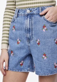 Pantalones cortos de denim azul con bordado floral en rojo y rosa, que presentan una cintura alta, bolsillos frontales y un corte clásico.