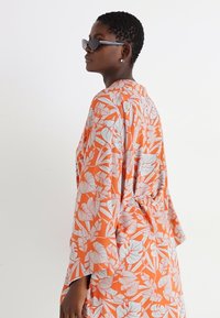 Kimono floral en tissu orange vif, avec des motifs de feuilles blanches, des manches longues et une taille nouée pour un ajustement décontracté.