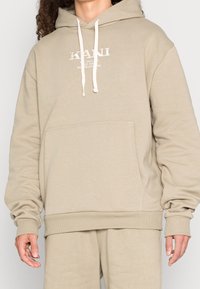 Béžová mikina s kapucí z bavlněné směsi. Má přední kapsu, nastavitelnou kapuci se stahovací šňůrkou a bílý vyšívaný logo na hrudi.