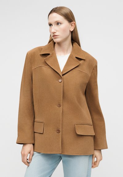 Γυναίκα με καφέ παλτό με κουμπιά, μεγάλους γιακάδες και τσέπες με καπάκια, που φοράει ανοιχτόχρωμο μπλε τζιν, κοιτάζοντας προς το πλάι σε απλό φόντο.