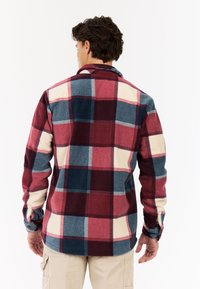 Man die met zijn rug naar de camera staat en een rood, blauw en crêmekleurig groot geruite fleecejack en beige cargobroek draagt.