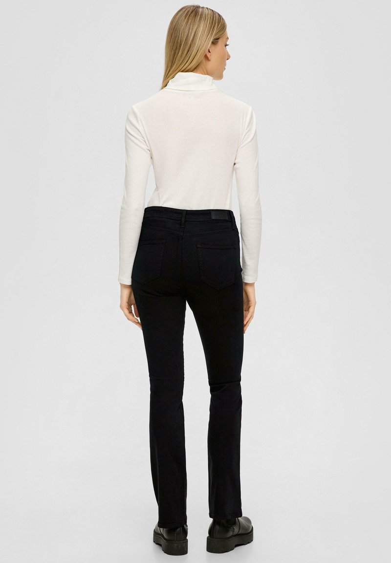 Schwarze Schlagjeans mit einer mittelhohen Taille, kombiniert mit einem engen weißen Rollkragenpullover. Die Jeans haben standardmäßige Gesäßtaschen und minimale Details.