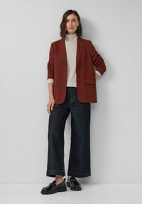 Vrouw staat tegen een lichte muur gekleed in een roestkleurige blazer, lichtgrijze coltrui, donkere wijde jeans en zwarte instappers.