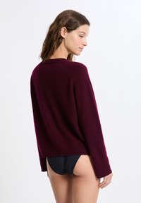 Frau mit braunen Haaren, die einen langärmeligen burgunderfarbenen Pullover und schwarze Bikinihosen trägt, seitlich vor einem weißen Hintergrund stehend.