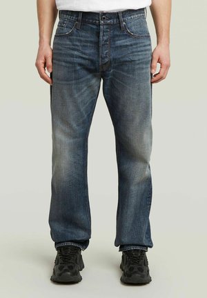Homme portant un jean droit bleu délavé et des baskets noires, debout devant un fond clair uni, les mains détendues le long du corps.