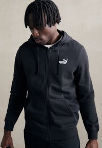 Sort zip-up hoodie lavet af blødt stof med en kengurulomme, der har et hvidt Puma-logo på venstre bryst og justerbar snoretræk-hætte.