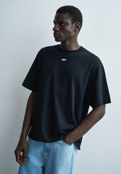 Jeune homme portant un t-shirt noir oversize avec le texte "OFF", jean bleu clair, debout avec une main dans la poche contre un mur uni.