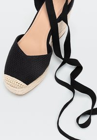 Espadrille noire avec tige en tissu tissé, bout rond et plateforme en jute naturel. Elle dispose de longues lanières noires pour les liens de cheville.