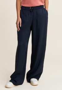 Pantalons larges bleu marine en tissu doux, avec une taille élastique dotée d'un cordon de serrage et des poches latérales. Associés à des baskets blanches.