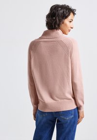 Suéter rosa claro con un alto cuello acanalado y acentos texturizados en los hombros. Material suave, puños ajustados y un dobladillo acanalado. Llevado con jeans azules.