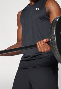 Maglietta atletica nera senza maniche con texture liscia e logo Under Armour sul petto, abbinata a pantaloni corti neri, mentre afferra un bilanciere.