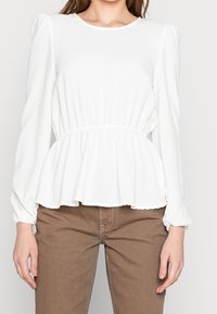 Weiße Peplum-Bluse mit langen Puffärmeln, weicher Textur, geraffter Taille und rundem Ausschnitt; getragen mit braunen Hosen.