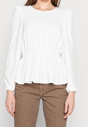 Blusa bianca a peplo con maniche lunghe a palloncino, morbida al tatto, con vita arricciata e scollatura rotonda; indossata con pantaloni marroni.