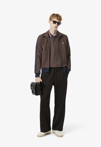Veste en tissu brun à motifs avec des accents bleu marine, pantalon noir, baskets blanches et sac noir ; modèle portant des lunettes de soleil.