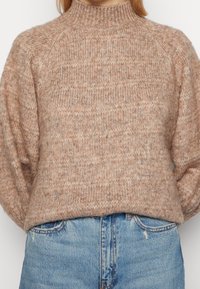 Pull en tricot beige clair avec un col montant, des manches raglan et un motif texturé. Associé à un jean en denim bleu clair.