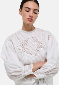 Blouse blanche avec une encolure en dentelle texturée, des manches bouffantes et des détails en dentelle raffinés. Présente un col montant et des poignets délicats.