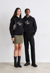 Crne dukserice imaju središnji bijeli logo i vezicu, u kombinaciji s maslinasto zelenom suknjom i crnim čizmama do koljena, na betonskoj pozadini.