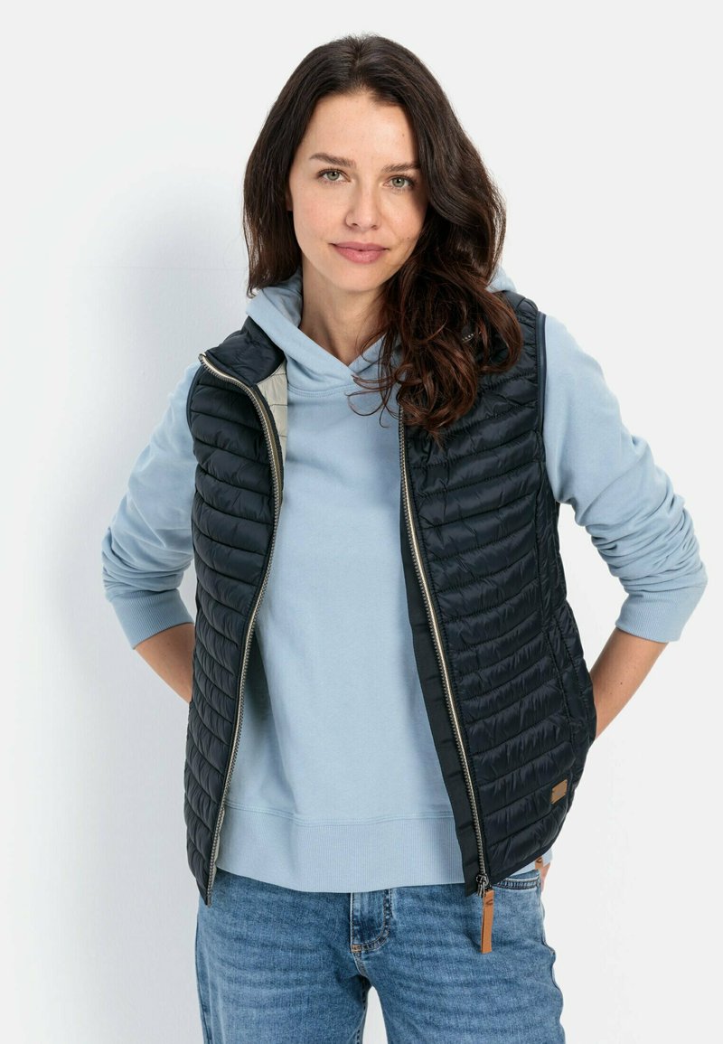 Zwarte gewatteerde vest met een hoge kraag, ritssluiting en een gestructureerd patroon, over een lichtblauwe hoodie. Model staat met de handen in de zakken.