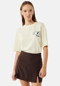 Gul bomulds T-shirt med Snoopy grafik og "Peanuts" tekst, parret med en mørkebrun miniskørt med en sideslids.