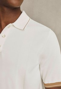 Valkoinen polo paita, jossa on ribattu pinta, kolmesta nappulasta koostuva etuosa ja kontrastiväri tummanruskea raita hihoissa.