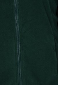 Giacca in fleece verde scuro con chiusura zip foderata, caratterizzata da una texture morbida e una finitura liscia. Nessun logo o motivo visibile.