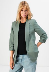 Mladá žena s blond vlasy, oblečená v sázavě zeleném saku, černém topu a světle modrých džínách, na čistém bílém pozadí.