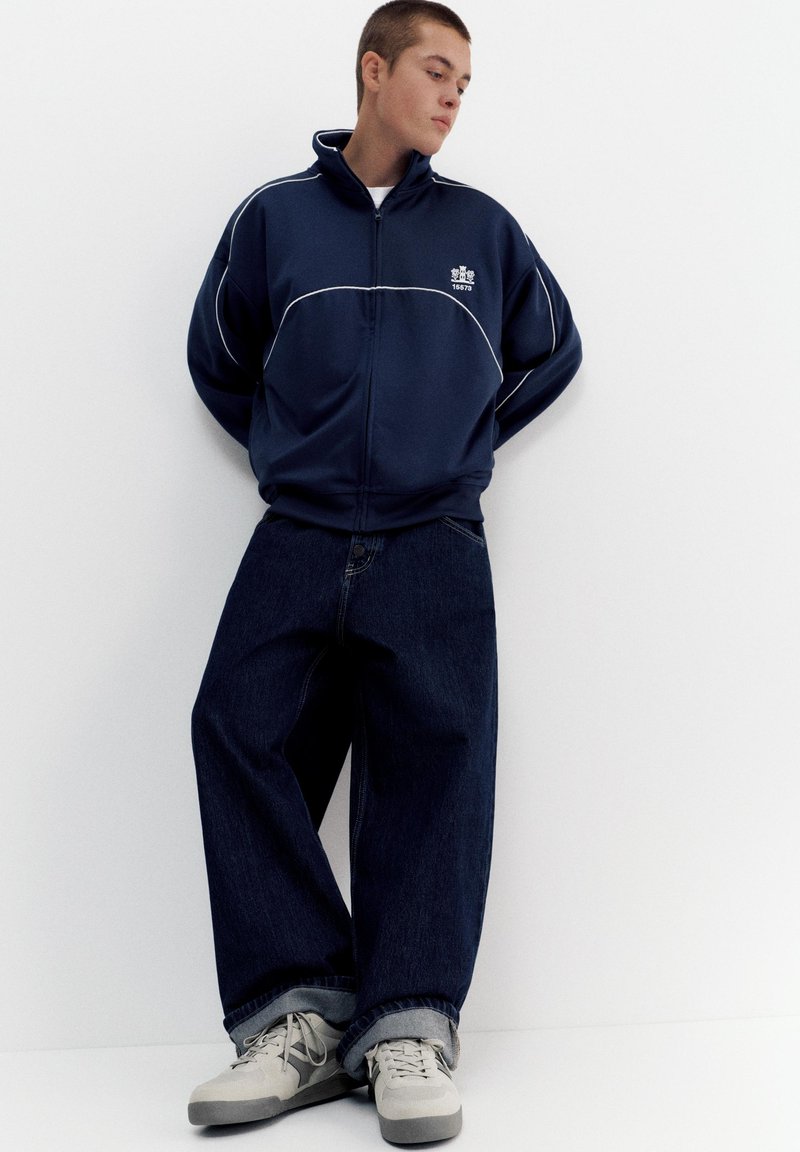 Jeune homme debout contre un mur blanc, portant une veste zippée bleu marine, un jean large foncé et des baskets grises, regardant sur le côté avec les mains derrière le dos.