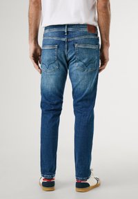 Jeans de denim azul de corte ajustado con un lavado desvaído, cinco bolsillos y detalles de costura visibles. Parche de cuero en la cinturilla trasera.